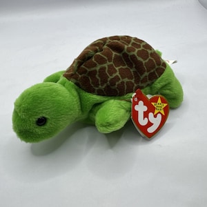 Peut inclure: Une peluche verte en forme de tortue avec une carapace marron à motifs. La tortue porte une étiquette rouge en forme de cœur avec le texte "Beanie Baby" et le logo "ty".