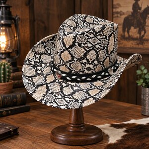 Pode incluir: Um chapéu de cowboy com um padrão de pele de cobra em bege, preto e branco. O chapéu inclui uma faixa preta com um acabamento trançado branco e detalhes circulares prateados. A aba é curvada para cima e a copa tem um vinco central.