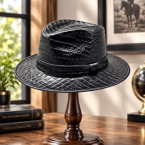 May include: A black fedora hat featuring a textured crocodile skin pattern across the crown and brim. The hat includes a matching black band encircling the base of the crown, providing a uniform appearance with a structured shape and a wide brim.