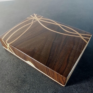 May include: A rectangular wooden box with a dark brown finish and inlaid gold-colored decorative lines. The box has a light-colored border and a smooth, polished surface. The design is geometric and elegant.