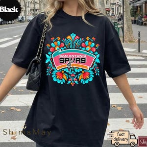 May include: Black t-shirt with a vibrant floral design and the words "San Antonio Spurs" in a pink and white banner. The shirt has a crew neck and short sleeves. The design includes teal, orange, and white accents.