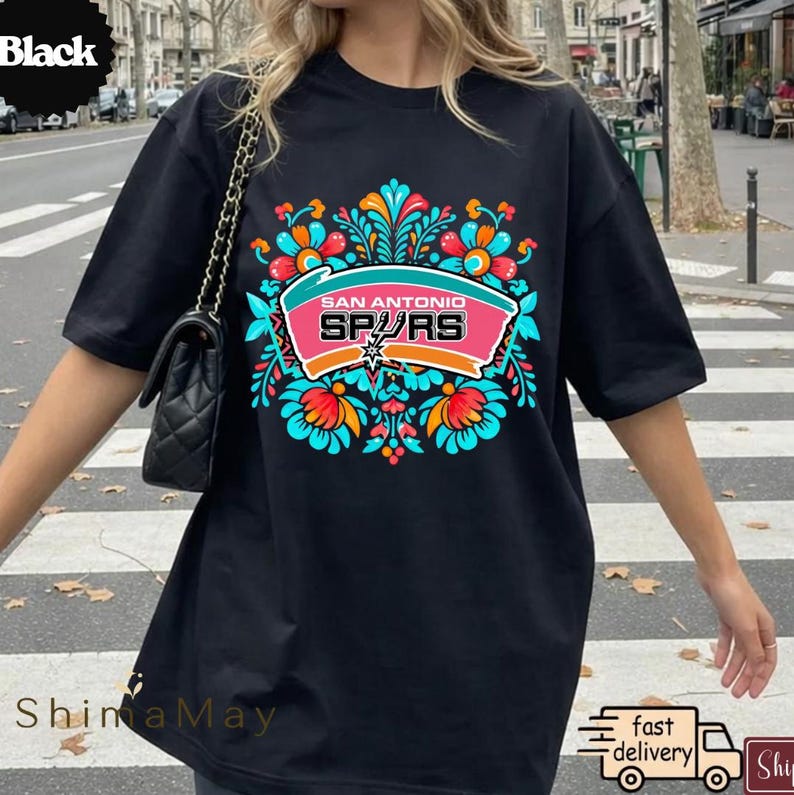 May include: Black t-shirt with a vibrant floral design and the words "San Antonio Spurs" in a pink and white banner. The shirt has a crew neck and short sleeves. The design includes teal, orange, and white accents.