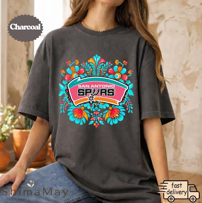 May include: A charcoal-coloured t-shirt with a vibrant floral design. The design includes the words "San Antonio Spurs" in a pink and teal banner, surrounded by orange, blue, and red floral elements. A casual wear item.
