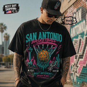 May include: Black t-shirt with a San Antonio Basketball graphic. The design features a basketball going through a net, city skyline, and the text "SAN ANTONIO BASKETBALL". Includes the text "EST. 1973 BUILT DIFFERENT DRIVEN TO WIN".