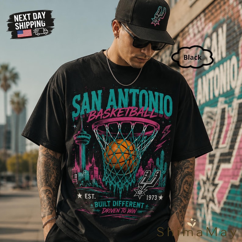 May include: Black t-shirt with a San Antonio Basketball graphic. The design features a basketball going through a net, city skyline, and the text "SAN ANTONIO BASKETBALL". Includes the text "EST. 1973 BUILT DIFFERENT DRIVEN TO WIN".