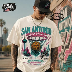 May include: Ivory-colored t-shirt featuring a San Antonio basketball design. The graphic includes a basketball going through a net, city skyline, and text that reads "SAN ANTONIO BASKETBALL" and "BUILT DIFFERENT". The shirt also has the text "EST. 1973".