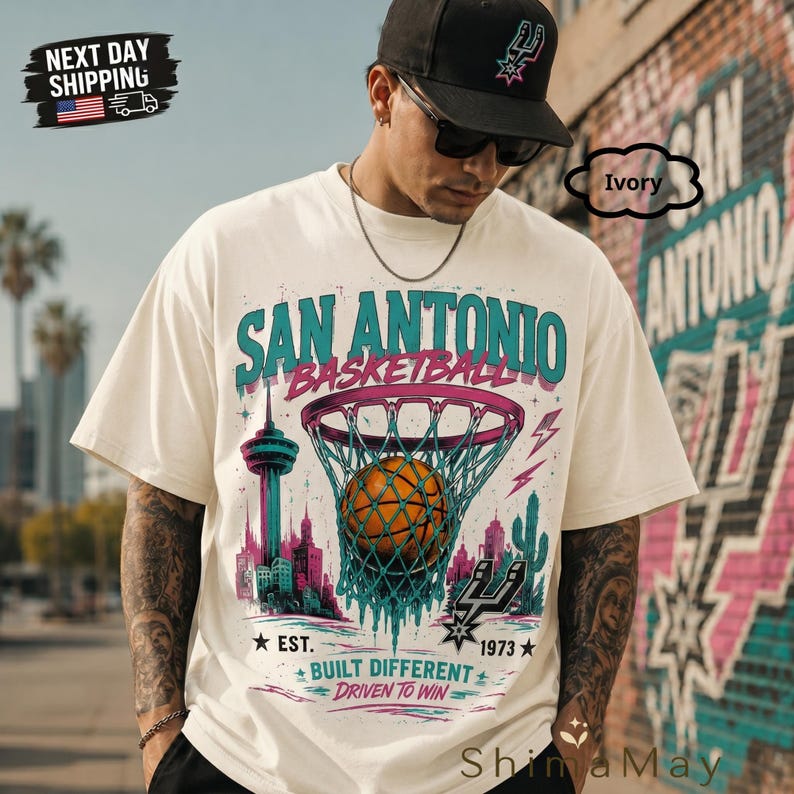 May include: Ivory-colored t-shirt featuring a San Antonio basketball design. The graphic includes a basketball going through a net, city skyline, and text that reads "SAN ANTONIO BASKETBALL" and "BUILT DIFFERENT". The shirt also has the text "EST. 1973".