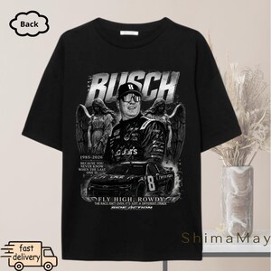 以下が含まれることがあります： レーサー、レーシングカー、天使の姿をグレースケールで描いた黒いTシャツ。「BUSCH」の文字が上部にあり、「1985-2026」などの文字が下にあります。Tシャツはクルーネックで半袖です。