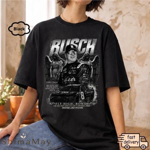 以下が含まれることがあります： 黒色のグラフィックTシャツ。レースカーのドライバーの肖像画、「BUSCH」の文字、レースカーが描かれています。シャツには「1985-2026」と「FLY HIGH, ROWDY」の文字も含まれています。デザインはグレーと白の濃淡です。