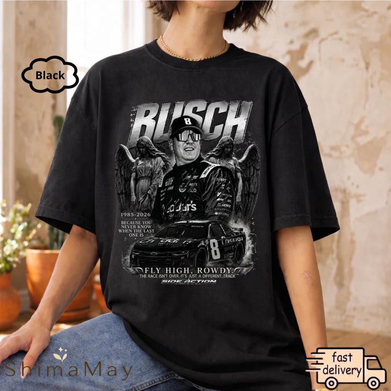 以下が含まれることがあります： 黒色のグラフィックTシャツ。レースカーのドライバーの肖像画、「BUSCH」の文字、レースカーが描かれています。シャツには「1985-2026」と「FLY HIGH, ROWDY」の文字も含まれています。デザインはグレーと白の濃淡です。