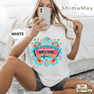 May include: White t-shirt with a vibrant floral design and the text "SAN ANTONIO SPURS" in a pink and teal banner. The shirt has a crew neck and short sleeves. The word "WHITE" is in a white bubble on the left side of the shirt.