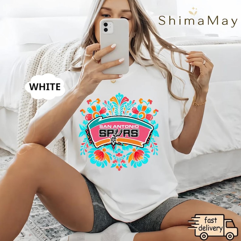 May include: White t-shirt with a vibrant floral design and the text "SAN ANTONIO SPURS" in a pink and teal banner. The shirt has a crew neck and short sleeves. The word "WHITE" is in a white bubble on the left side of the shirt.