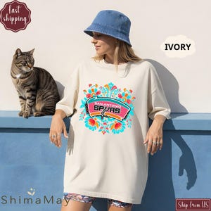 May include: An ivory-coloured oversized t-shirt with a colourful floral design and the text "San Antonio Spurs". The shirt is worn with a blue bucket hat. A tabby cat is sitting on a blue ledge next to the person wearing the shirt.