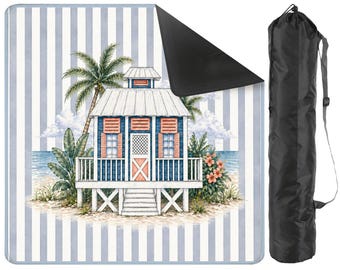 May include: A picnic blanket with a blue and white striped design, featuring a beach scene illustration. The image shows a beach house, palm trees, and flowers. A black carry bag is also visible.