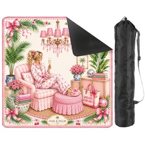 May include: A pink and white patterned beach blanket with a black underside, featuring a woman in pajamas, a pink armchair, and a chandelier. Includes a black carrying bag with a shoulder strap. The blanket has the text "GOOD DAYS & TAN LINES".