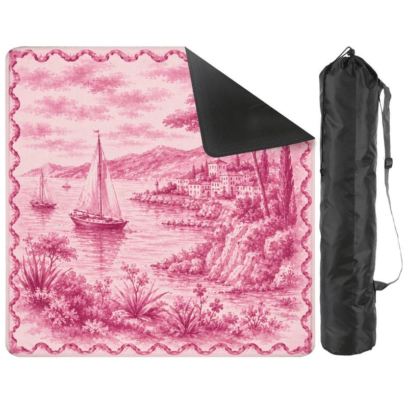 May include: A pink and white picnic blanket with a scenic illustration of sailboats on a lake, a village, and mountains. The blanket has a black underside and a decorative border. A black carrying bag is included.
