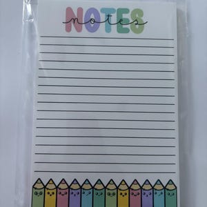 May include: A rectangular notepad in a clear plastic sleeve featuring the word NOTES in colorful bubble letters at the top, horizontal black lines for writing, and a row of colorful cartoon pencils with faces along the bottom edge.