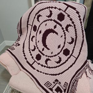May include: A light pink crochet blanket featuring a dark maroon circular design of moon phases and stars. The blanket has a geometric border pattern and fringe along the edges, showcasing a detailed tapestry-style stitch throughout the fabric.