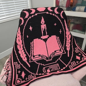 Puede incluir: Una manta de ganchillo negra y rosa con un diseño central de un libro abierto con una vela encendida encima. El borde muestra un patrón de fases lunares, estrellas y formas geométricas. La manta tiene flecos negros y rosas en el lateral.