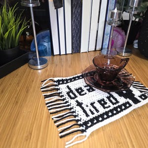 Może przedstawiać: Prostokątna szydełkowa podstawka z białym tłem, czarnym napisem "dead tired" i czarną obw&oacute;dką. Podstawka posiada frędzle po dw&oacute;ch stronach i służy jako podkładka pod ciemnofioletową szklaną filiżankę do herbaty ze spodkiem.