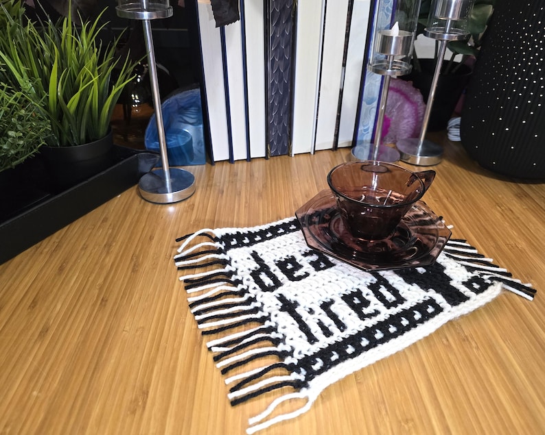 Może przedstawiać: Prostokątna szydełkowa podstawka z białym tłem, czarnym napisem "dead tired" i czarną obw&oacute;dką. Podstawka posiada frędzle po dw&oacute;ch stronach i służy jako podkładka pod ciemnofioletową szklaną filiżankę do herbaty ze spodkiem.