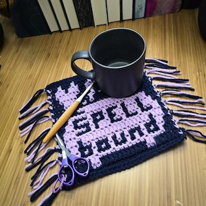 Puede incluir: Una taza morada oscura se encuentra sobre un posavasos de ganchillo hecho a mano con las palabras "SPELL BOUND" en morado claro. Un ganchillo y unas tijeras moradas tambi&eacute;n est&aacute;n sobre el posavasos. El posavasos tiene un borde y flecos azul marino.