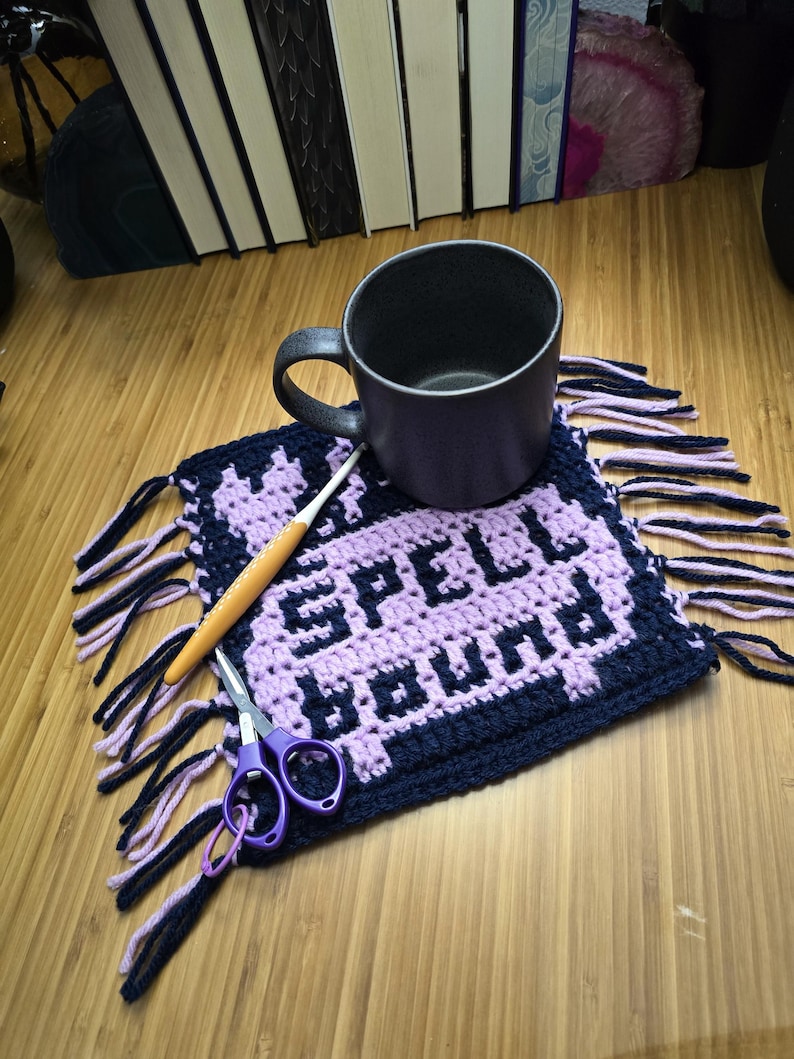 Puede incluir: Una taza morada oscura se encuentra sobre un posavasos de ganchillo hecho a mano con las palabras "SPELL BOUND" en morado claro. Un ganchillo y unas tijeras moradas tambi&eacute;n est&aacute;n sobre el posavasos. El posavasos tiene un borde y flecos azul marino.
