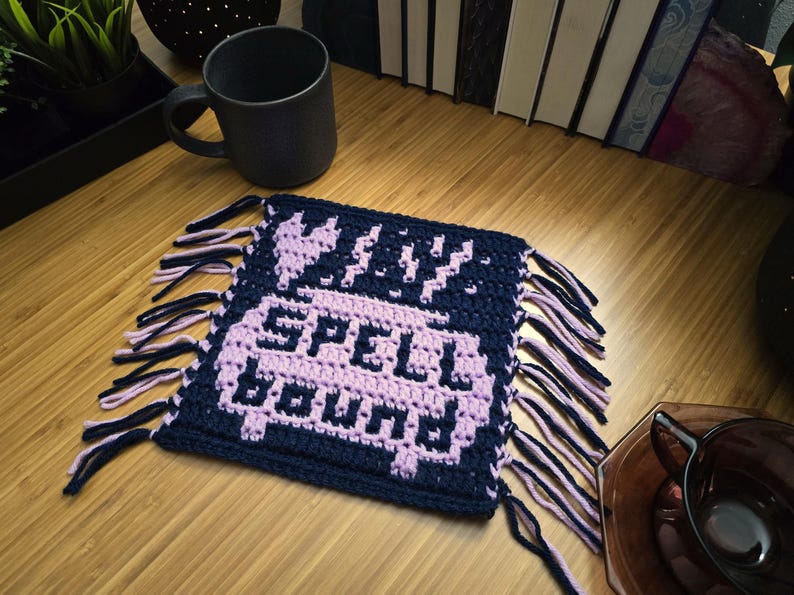Puede incluir: Un posavasos de ganchillo azul marino y lavanda con las palabras "WILY SPELL BOUND" en un dise&ntilde;o pixelado. El posavasos tiene flecos en los bordes y est&aacute; sobre una superficie de madera. Una taza negra est&aacute; en el fondo.