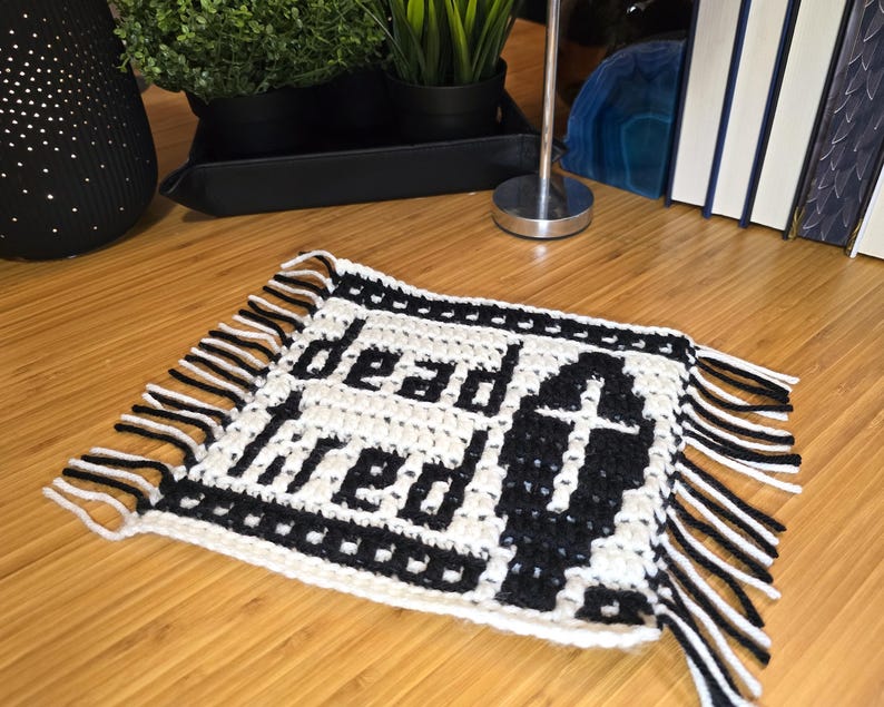 Może przedstawiać: Kwadratowa szydełkowa podstawka pod kubek w czarno-białej kolorystyce. Wz&oacute;r przedstawia napis "dead tired" pogrubioną czcionką oraz grafikę trumny z krzyżem z przodu. Krawędzie wykończone są czarno-białymi frędzlami.