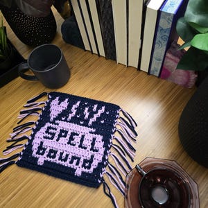 Puede incluir: Un posavasos de ganchillo azul marino y lavanda con las palabras "SPELL BOUND" en lavanda. El posavasos tiene flecos en los bordes. Una taza negra y una pila de libros est&aacute;n en el fondo.