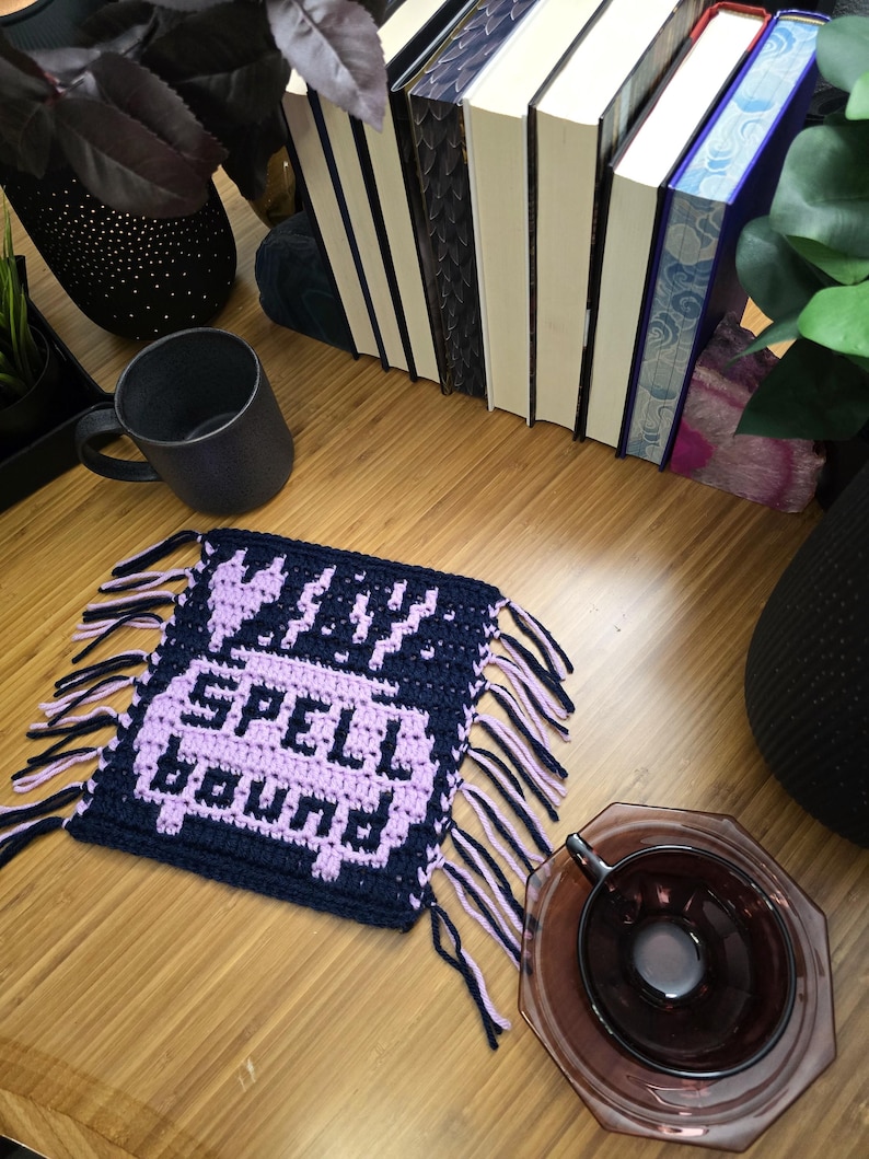 Puede incluir: Un posavasos de ganchillo azul marino y lavanda con las palabras "SPELL BOUND" en lavanda. El posavasos tiene flecos en los bordes. Una taza negra y una pila de libros est&aacute;n en el fondo.