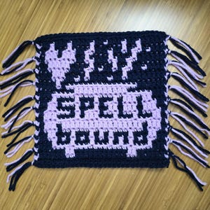 Puede incluir: Una pieza de ganchillo hecha a mano en azul marino y lavanda, con las palabras "SPELL BOUND" en letras pixeladas lavanda. El dise&ntilde;o incluye un coraz&oacute;n y rayos. Los bordes est&aacute;n acabados con flecos.