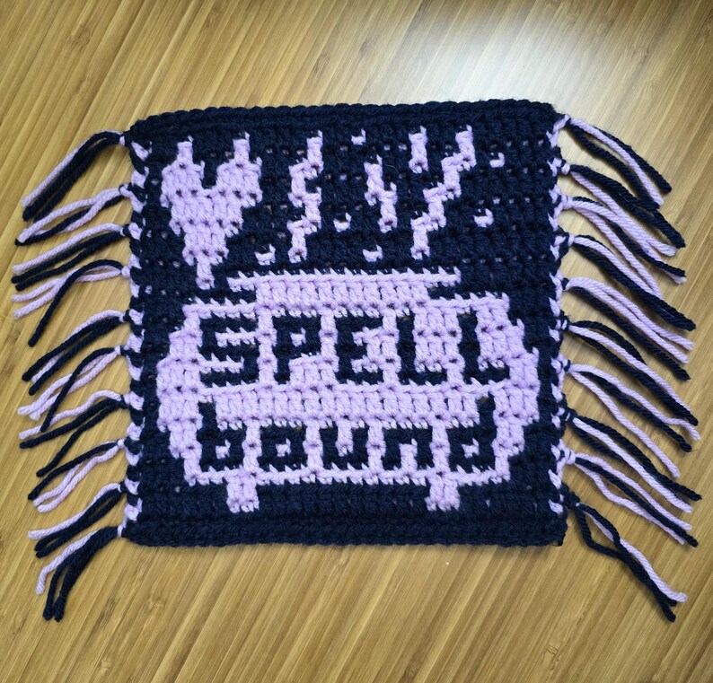 Puede incluir: Una pieza de ganchillo hecha a mano en azul marino y lavanda, con las palabras "SPELL BOUND" en letras pixeladas lavanda. El dise&ntilde;o incluye un coraz&oacute;n y rayos. Los bordes est&aacute;n acabados con flecos.