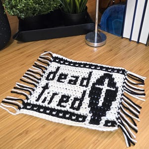 Może przedstawiać: Prostokątna szydełkowa podstawka pod kubek w czarno-białej kolorystyce. Wz&oacute;r przedstawia napis "dead tired" pogrubioną czcionką oraz grafikę trumny z krzyżem. Całość wykończona jest ozdobnymi frędzlami po obu stronach przedmiotu.