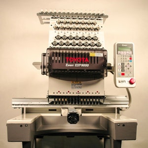 Puede incluir: Una m&aacute;quina de bordar Toyota Expert EST 9000 blanca con un panel de control negro y un bot&oacute;n rojo. La m&aacute;quina tiene un gran n&uacute;mero de agujas y una cubierta de pl&aacute;stico transparente sobre el &aacute;rea de las agujas. La m&aacute;quina est&aacute; sobre un soporte blanco con una base gris.