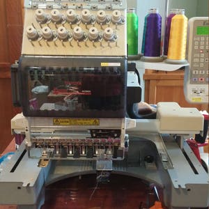 May include: A white and gray industrial embroidery machine with multiple spools of thread in green, purple, and yellow. The machine has a digital display and a warning label that reads "Caution".