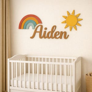May include: Wooden wall decorations including a rainbow, a yellow sun, and the name "Aiden" in a cursive font. The rainbow has layers of red, orange, teal, and blue. The sun is yellow. The name is in a warm brown colour. A white cot is in the foreground.