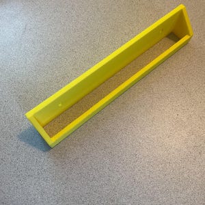 May include: A bright yellow rectangular shelf with a raised front and back. The shelf is made of a solid material and has two small holes on the top edge. The shelf is designed to be mounted on a wall.