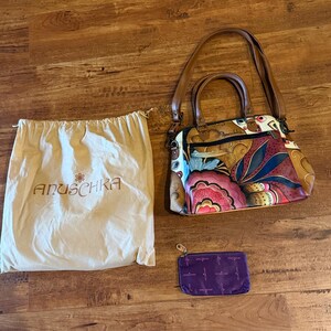 Peut inclure: Un sac à main multicolore à motif floral, doté de poignées et d'une bandoulière en cuir marron, accompagné d'un sac de protection beige à cordon avec le logo Anuschka et d'une petite pochette rectangulaire zippée violette à logo répété.