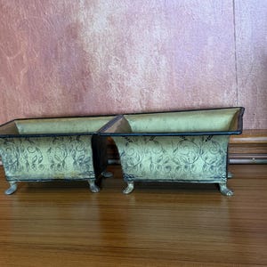 May include: Two rectangular metal planters with a distressed gold finish and black painted vine patterns. Each planter features a flared rim, a dark border, and four small, ornate feet that elevate the base from the surface.