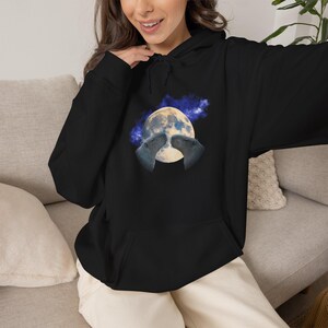 May include: A black hooded sweatshirt featuring a graphic print on the chest. The design displays two capybaras facing each other in front of a large, detailed full moon set against a dark blue, starry, and nebulous cosmic background.