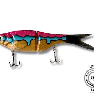 May include: A custom fishing lure designed to resemble a donut. The lure features a pink top with sprinkles, blue icing, and a golden-brown body. It has two treble hooks and a black tail. The logo "LETR Baits" is visible.