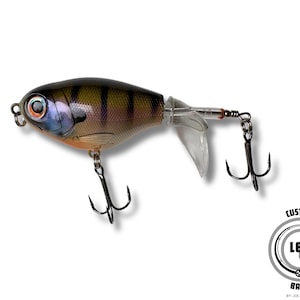 May include: A detailed fishing lure with a realistic design, featuring a brown and yellow body with black stripes. It has a clear plastic tail and two treble hooks. The lure is designed to mimic a small fish.