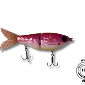 May include: A pink and gold fishing lure shaped like a fish, with two treble hooks and a brown tail fin. The lure has a black eye and is segmented in the middle. The logo for L.E.T.R. Baits is in the bottom right corner.