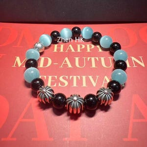 Könnte beinhalten: Ein Armband mit abwechselnd hellblauen und schwarzen kugelförmigen Perlen. Das Armband enthält auch silberfarbene dekorative Perlen. Der Hintergrund ist eine rote Oberfläche mit dem Text "Zhen HK HAPPY MID-AUTUMN FESTIVAL".