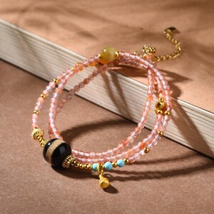May include: A multi-strand bracelet with pink and gold beads, a black and white patterned bead, and gold accents. The bracelet includes a small gold charm and turquoise beads. The jewellery is displayed on a neutral background.