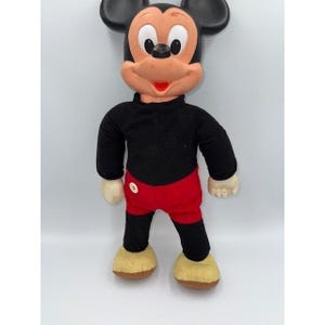 Könnte beinhalten: Eine Vintage-Plüschpuppe von Mickey Mouse. Die Puppe hat einen schwarzen Kopf und Ohren, einen roten Mund und ein schwarzes Hemd. Sie trägt rote Shorts, schwarze Beine und gelbe Schuhe. Die Puppe steht aufrecht vor einem weißen Hintergrund.