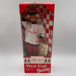 May include: A West End Barbie doll in a red and white box. The doll wears a white long-sleeved shirt with the red text Hamleys, a black and white hat, and red pants. The box features a red and white checkered pattern with small figures and the text Hamleys, The Finest Toy Shop In The World, and West End Barbie.