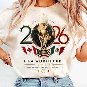 May include: A cream-colored t-shirt featuring a graphic of the 2026 FIFA World Cup. The design includes a gold trophy, a soccer ball, and the flags of Canada, the United States, and Mexico. Text reads FIFA WORLD CUP 2026 THREE NATIONS. ONE WORLD. ONE GOAL.