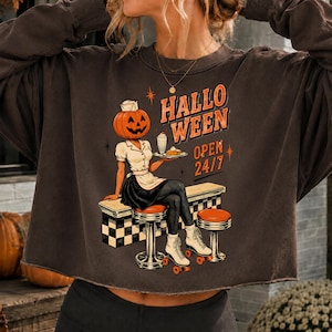 Könnte beinhalten: Ein dunkelbraunes Langarm-Sweatshirt mit einer Grafik einer Figur mit Kürbiskopf in Diner-Uniform auf Rollschuhen. Der Text lautet HALLOWEEN OPEN 24/7. Die Figur hält ein Tablett mit einem Milchshake und einem Stück Kuchen an einer Diner-Theke.
